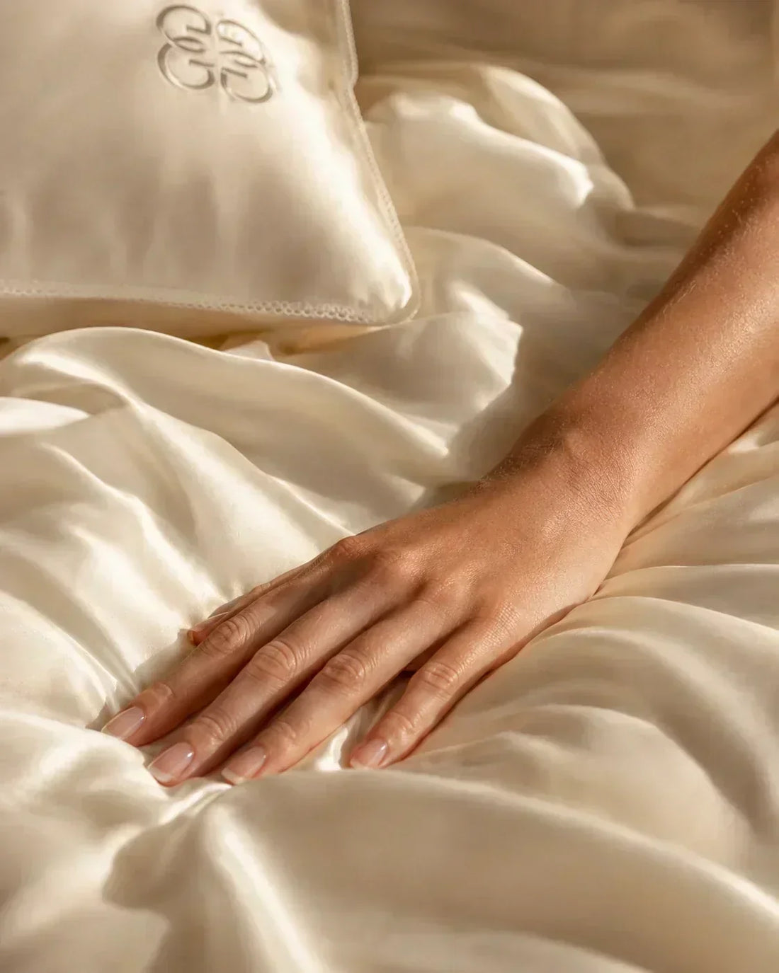 Hand resting on luxurious ivory 6A-grade mulberry silk bedding in soft natural light.