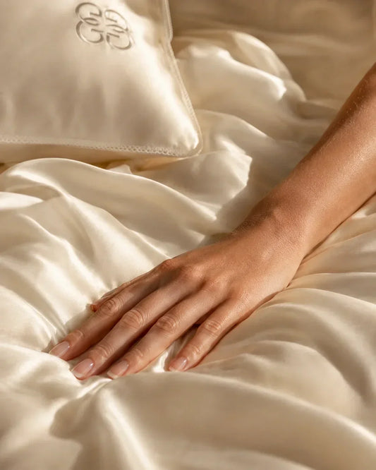 Hand resting on luxurious ivory 6A-grade mulberry silk bedding in soft natural light.